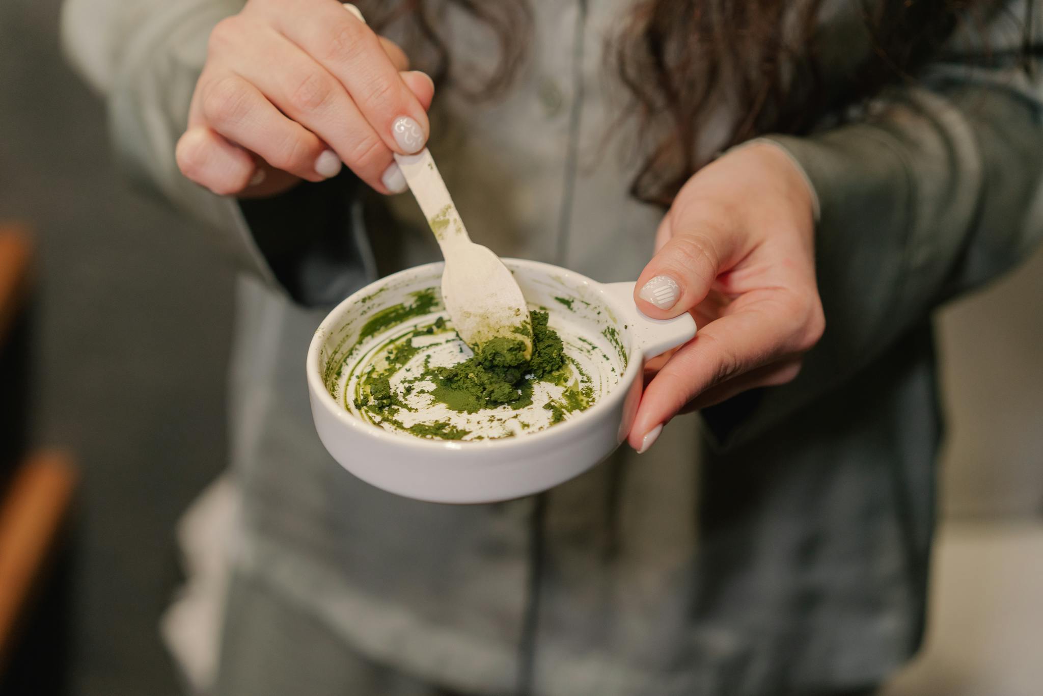Close-up of a woman preparing a green paste in a ceramic bowl, focusing on hands and texture.